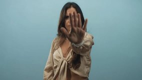 Woman with hand raised palm forward stop gesture in studio, long brown hair and tied blouse against light blue backdrop; defiance boundary. - Powered by Shutterstock - Get 15% off with code: PIKWIZARD15
