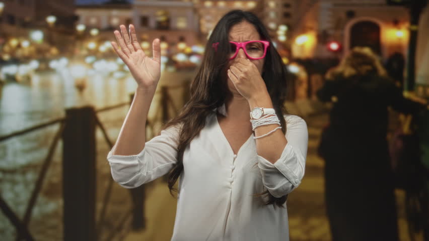Woman pinching nose on city street by riverside railing wearing pink glasses and white blouse, hand raised; urban discomfort.