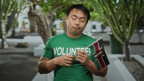 Young asian man in a green volunteer shirt holds a kenyan flag in an urban outdoor setting with trees lining the background, expressing a look of curiosity. - Powered by Shutterstock - Get 15% off with code: PIKWIZARD15