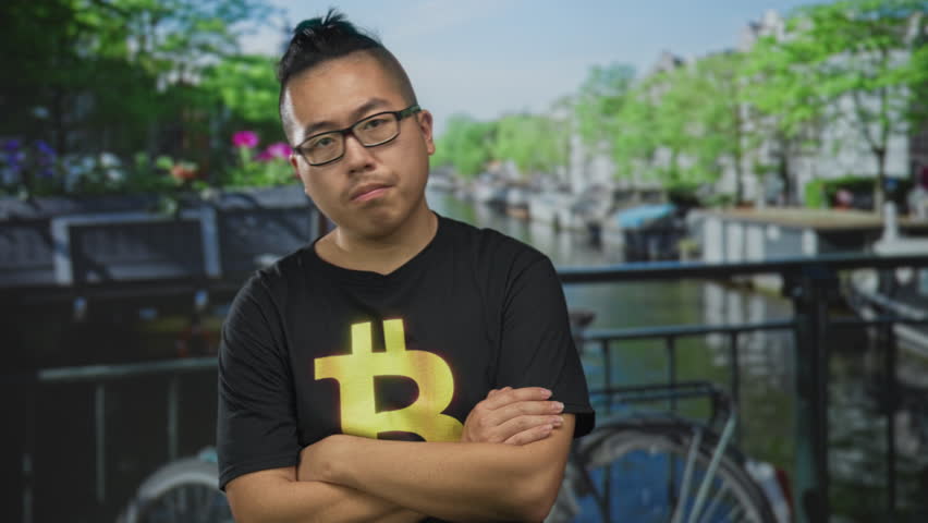Man in black bitcoin t shirt with arms crossed on street by canal and bicycle in amsterdam; crypto confidence investment.