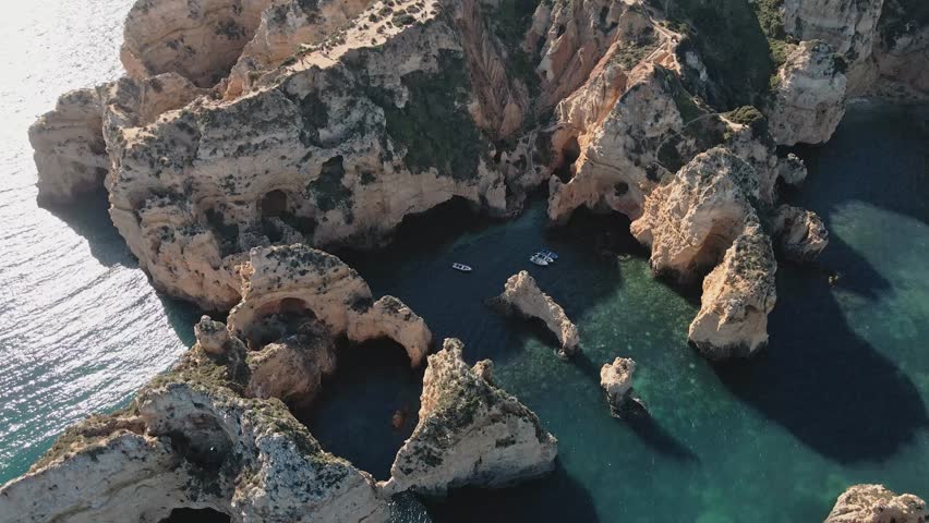 Aerial drone performs a helix orbit, circling above the limestone arches, sea stacks, and small boats nestled in the turquoise waters of Ponta da Piedade near Lagos, Portugal.