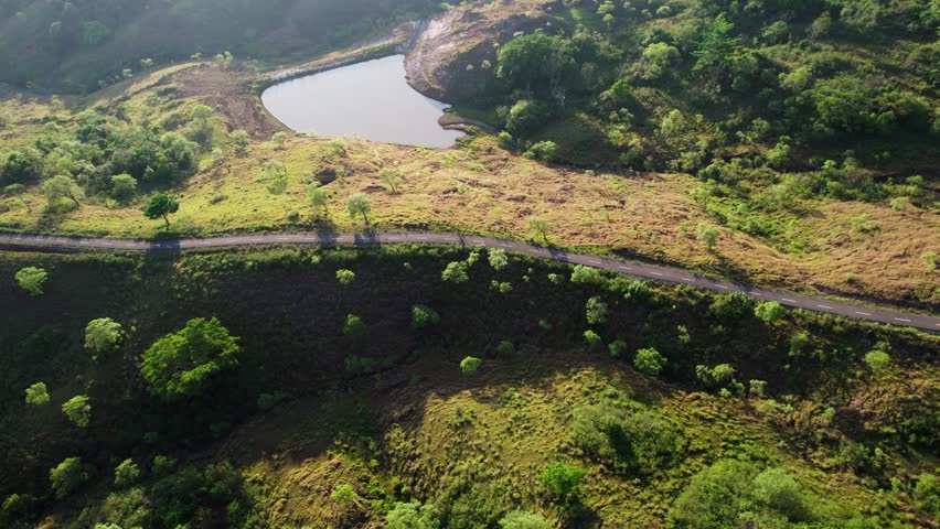 Aerial drone footage of an empty asphalt road winding past a small lake or reservoir. The remote landscape features green grassy hills and trees, illuminated by morning sunlight in Indonesia.