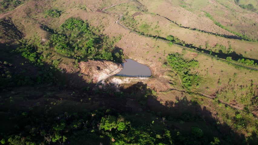 A high-angle aerial drone shot capturing a small lake nestled amidst rolling hills. Strong morning sunlight creates dramatic shadows across the green and brown landscape. Serene nature scene.