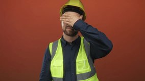 Man builder in yellow hardhat and high visibility vest covering eyes with hand in orange studio; uncertainty concealment. - Powered by Shutterstock - Get 15% off with code: PIKWIZARD15