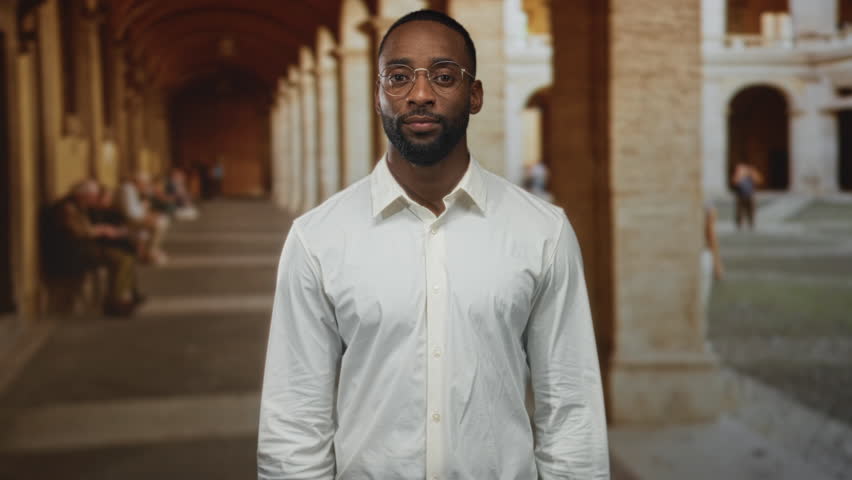 Man making air quotes with hands in building colonnade of old town street, wearing white shirt and round glasses; skepticism ironic wit.