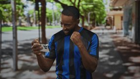 Man holds smartphone in hand and fist pumps on street wearing blue soccer jersey and braces while smiling; team pride. - Powered by Shutterstock - Get 15% off with code: PIKWIZARD15