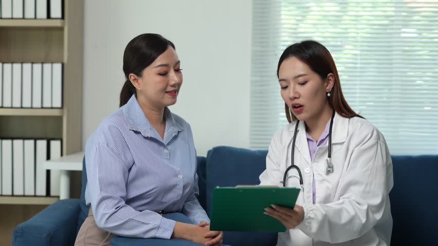 Professional female doctor in white coat with stethoscope is sitting on sofa and talking to patient, filling out medical history on clipboard
