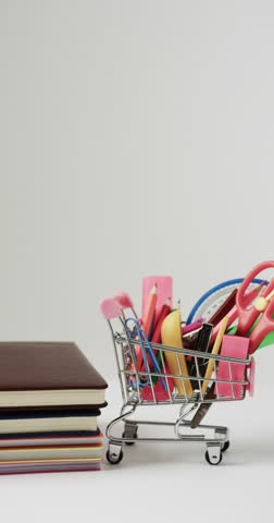 Vertical video of close up of shopping trolley with school items and books on grey background. Reading, creativity, learning, school and education concept.