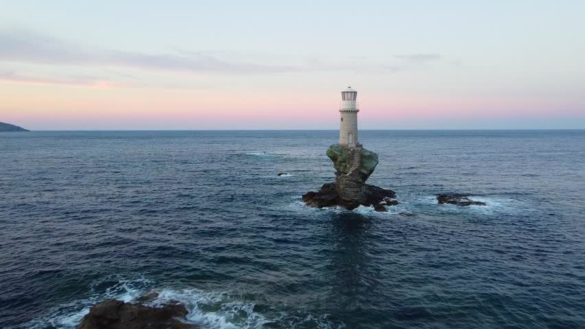 Tourlitis Lighthouse with spiral staircase on a rocky islet from the port of Chora at sunset, on Greek island of Andros, Aegean Sea, Drone shot