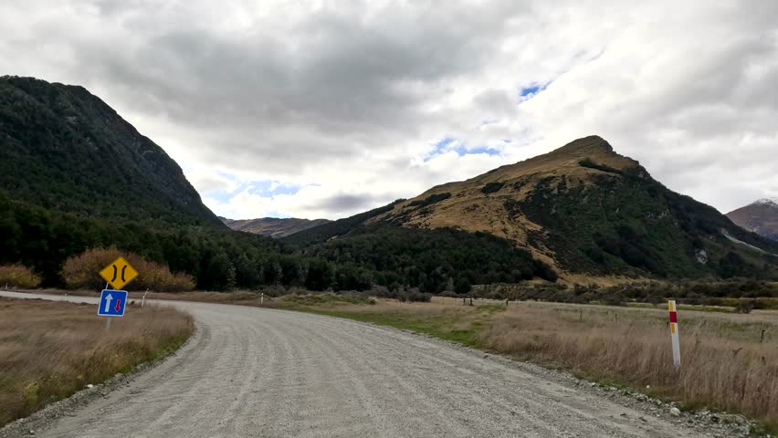 Vehicle travels gravel road through mountainous landscape, approaching bridge under cloudy daylight, steady forward motion