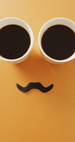 Vertical video of two cups of coffee and black false moustache on orange background with copy space. Father