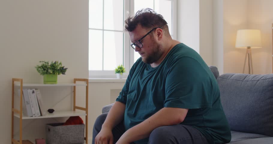 Tired fat overweight obese man holds forehead on couch. Male person with obesity shows headache, fatigue, stress, pain, migraine, exhaustion, and worry while sitting on sofa at home