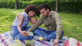 Young diverse couple enjoying a romantic date, sitting on a blanket on the green grass, taking funny selfies together with a smartphone - Powered by Shutterstock - Get 15% off with code: PIKWIZARD15