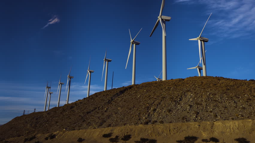 First-person passenger view looking up through a car window at wind turbines slowly turning atop a rocky ridge in the Mojave Desert of Southern California under a blue sky with wispy clouds.