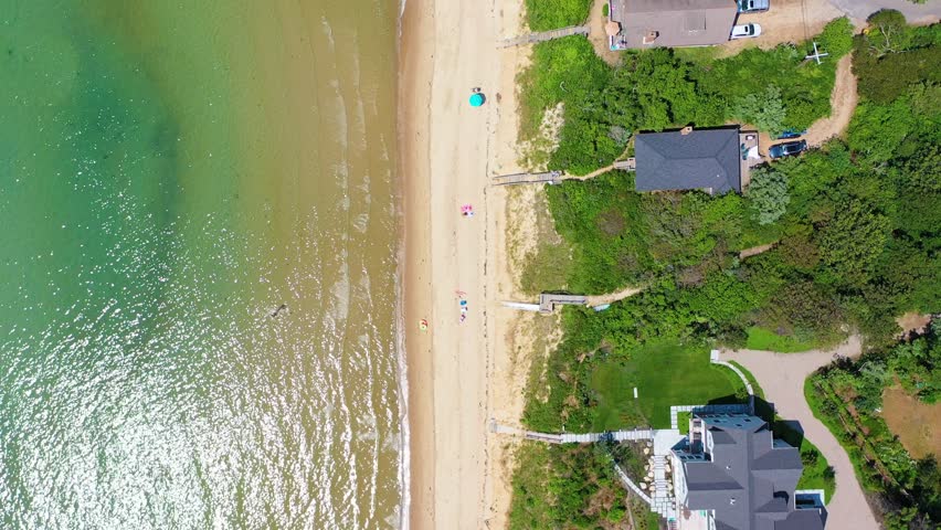 Cape Cod aerial drone view shows classic New England homes perched above sandy shoreline, stairways reaching to the beach, and ocean waters glistening in afternoon sun.