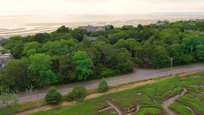 Aerial view of beachfront houses overlooking sandy shoreline and protective stone walls, framed by green dunes and wide marshland stretching across the horizon under a summer sky.