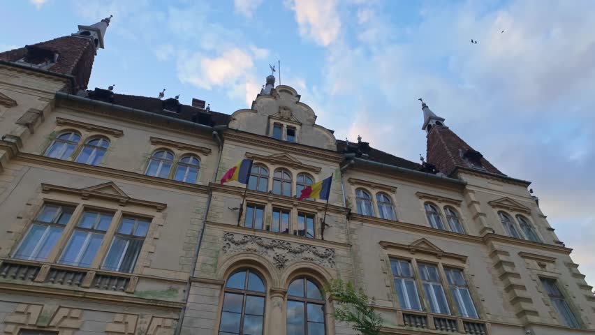 Rear facade of the Sighișoara City Hall, an elegant neo-Renaissance building with twin towers, arched windows and Romanian flags, seen under a soft evening sky.