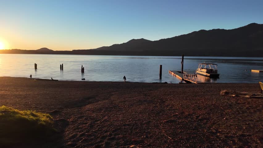 4K smooth stabilized footage of sunset at Quinault Lake in Olympic National Park, USA, showing a child’s silhouette playing on sandy beach near a dock and boat, golden evening light over calm water
