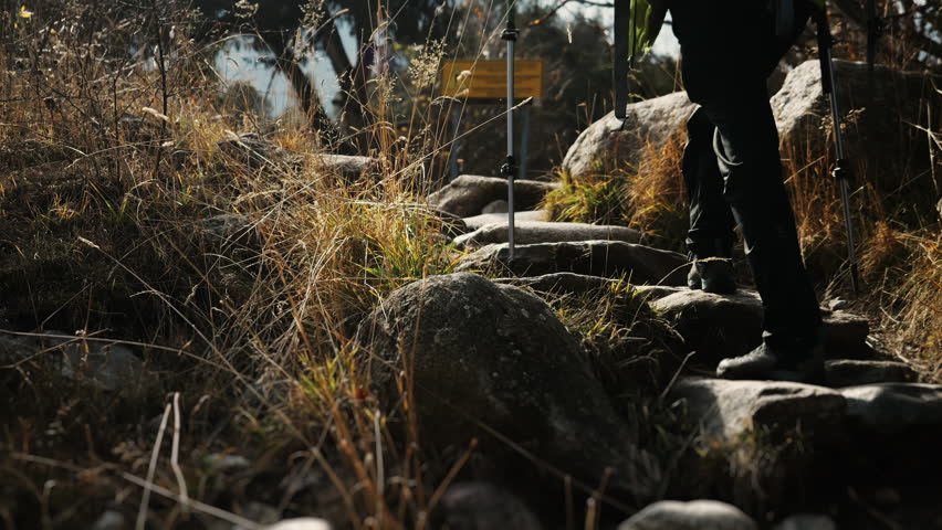 Hiker climbing a rocky mountain trail with a large backpack and trekking poles during an outdoor trek, surrounded by dry grass and natural terrain under sunlight.