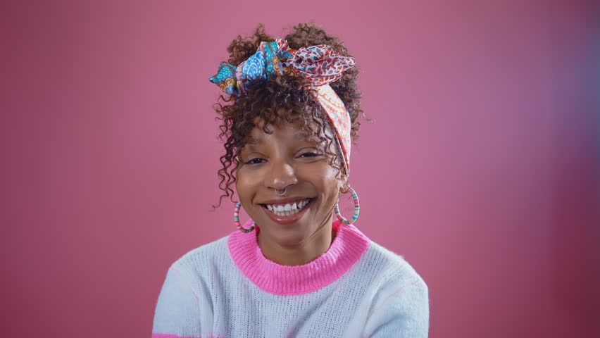 Studio shot of a cheerful young black woman with curly hair and colorful headscarf, wearing earrings and a light blue and pink sweater, smiling and looking away against a vibrant pink background