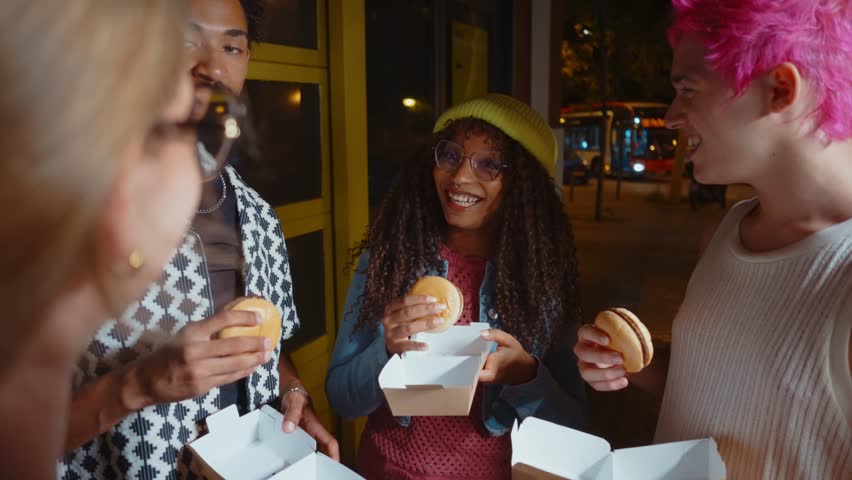 Young diverse friends smiling and laughing together while eating take-out food from boxes on a city street at night, enjoying their friendship and having a good time in a vibrant urban setting