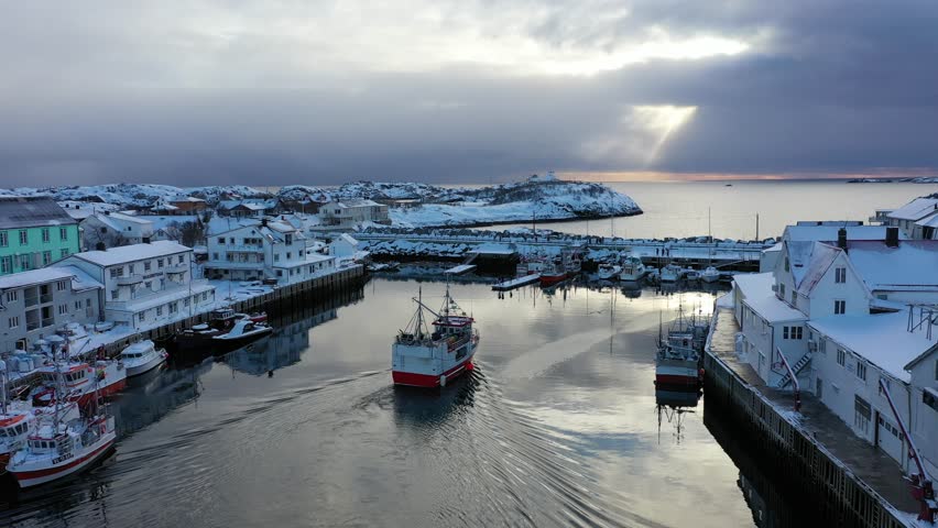Aerial view of Henningsvær harbor, with boats and snow-covered buildings under a cloudy sky, creating a serene winter scene, Henningsvær, Nordland, Norway.