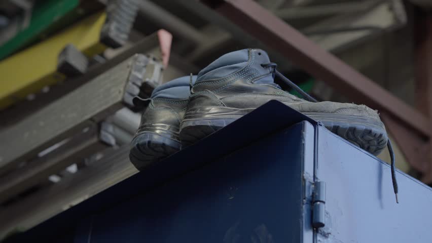 Low-angle close-up of a pair of worn work boots placed on top of a blue locker, with the factory roof visible in the background.
