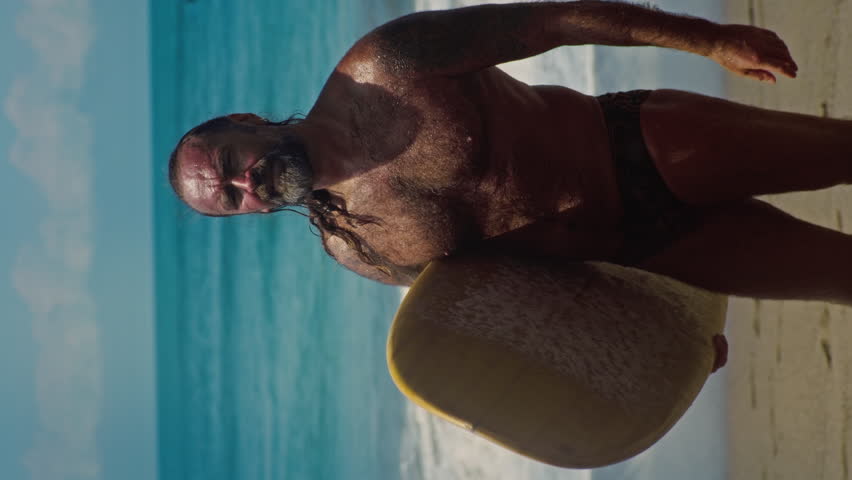 Vertical: senior having fun surfing at sunset time. Sporty bearded old man training with surfboard on the ocean beach. Elderly healthy male lifestyle, extreme sport concept. Active lifestyle, tropical