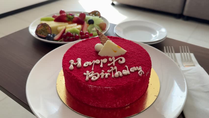 cake with the inscription "Happy Birthday" on a red background, decorated with white icing and a candle in the shape of a number. Next to the cake is a plate with fruits: pineapple, strawberry, kiwi, 