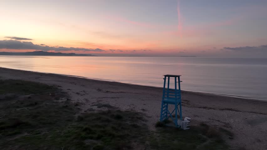 Serene sunrise over a calm sea with a blue lifeguard tower on the sandy beach. Aerial drone view of a tranquil morning coastline