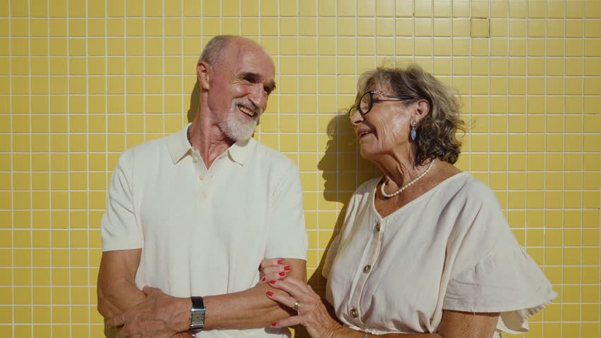 Happy senior couple sharing a joyful moment and laughing together while standing against a bright yellow tiled wall, enjoying their retirement and showcasing their affectionate, long-lasting love