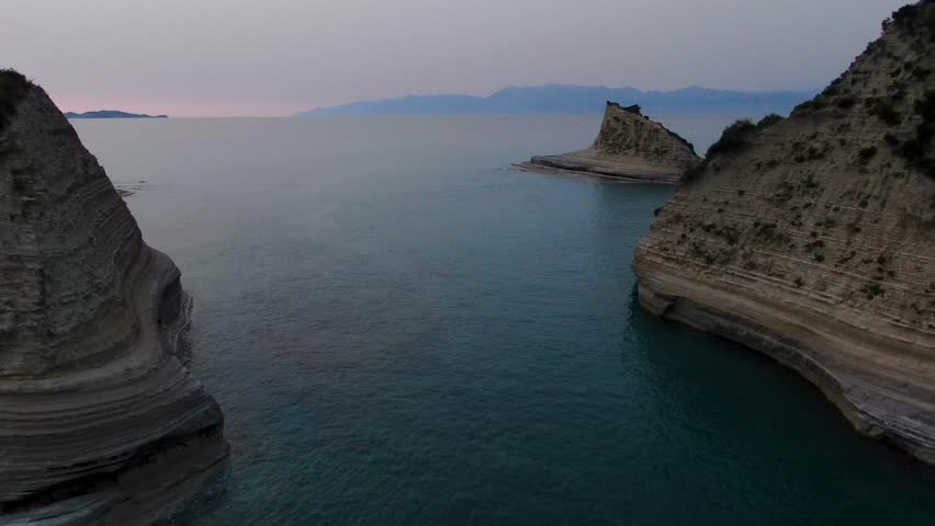 Cinematic drone flight through dramatic sea cliffs revealing a stunning rock formation in the ocean at dusk. Cape Drastis, Corfu
