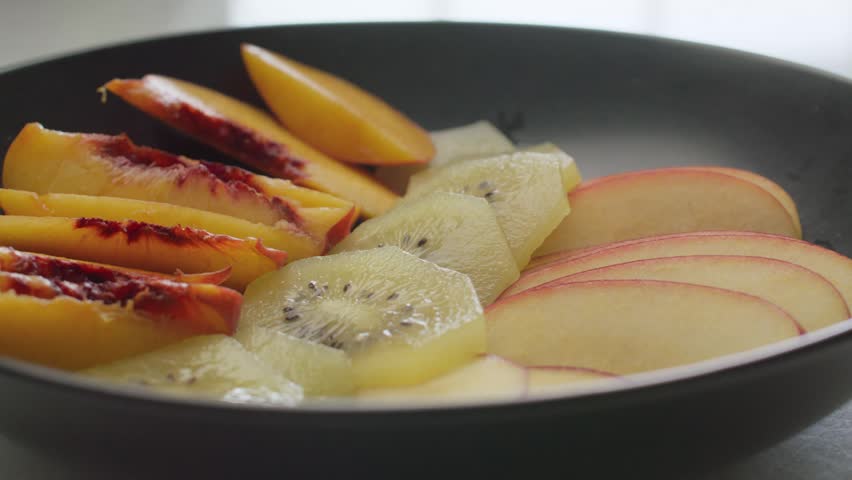 Close centered shot of a fruit salad in a black plate on a white table, with peach, kiwi and apple slices arranged in rows.