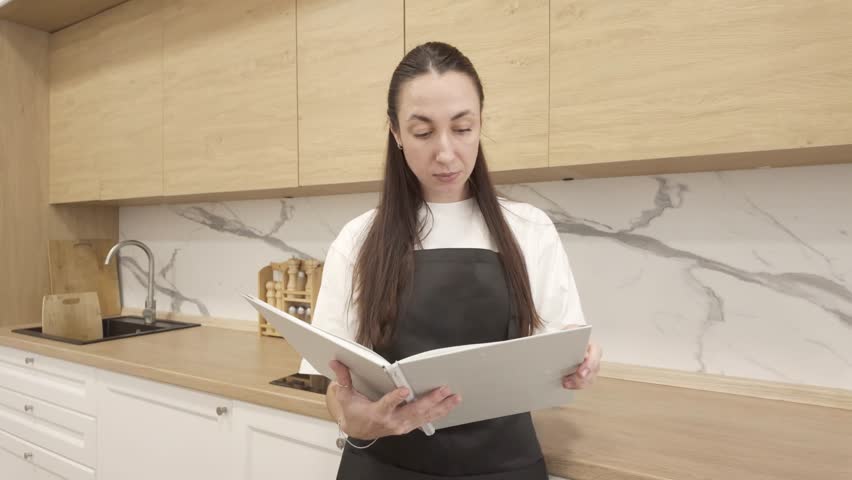 Young woman in black apron standing in modern kitchen and reading recipe book, preparing to cook and concentrating on new meal ideas at home