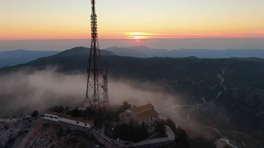 Aerial orbit of a mountaintop monastery and large communication tower on Mount Pantokrator, Corfu, Greece, at sunrise with fog in the valleys