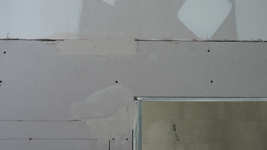 Close up of a worker using a metal spatula to apply plaster and fill joints between drywall panels during renovation in a new apartment. The process demonstrates accuracy, craftsmanship, and smooth