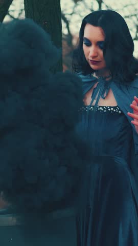 Medieval fantasy woman witch, hand waving casting spell. Magical black smoke from vat poison. lady queen blue velvet long vintage dress cape. Autumn nature dark gothic forest tree. vertical video crop
