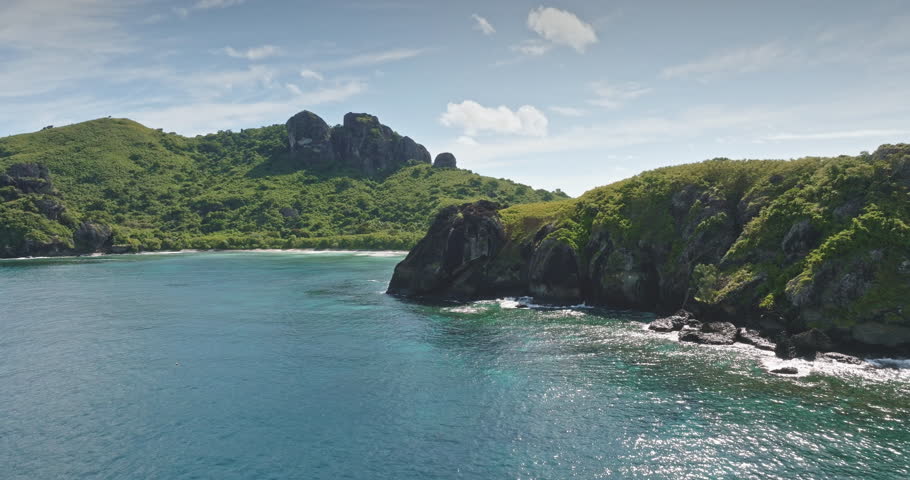 Fiji, Yaukuve Levu tropical island showing lush green mountains, rocky coastline, and white sand beach with turquoise and deep blue Pacific Ocean water under a light blue sky. Aerial view drone flight