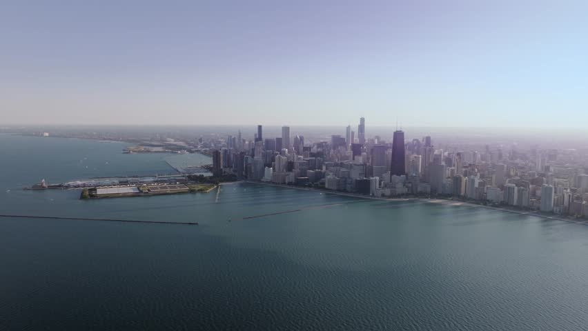 A high and wide, cinematic drone shot, captured far over Lake Michigan, slowly descending and tracking forward toward Navy Pier, and the sprawling metropolis and unique urban landscape of Chicago, IL.