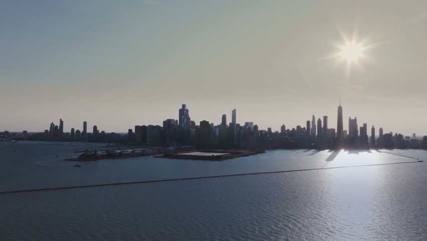 A wide, cinematic drone shot far over Lake Michigan, slowly rotating from left to right, as the setting sun casts a warm glow over the city skyline of towering skyscrapers of Chicago, IL.