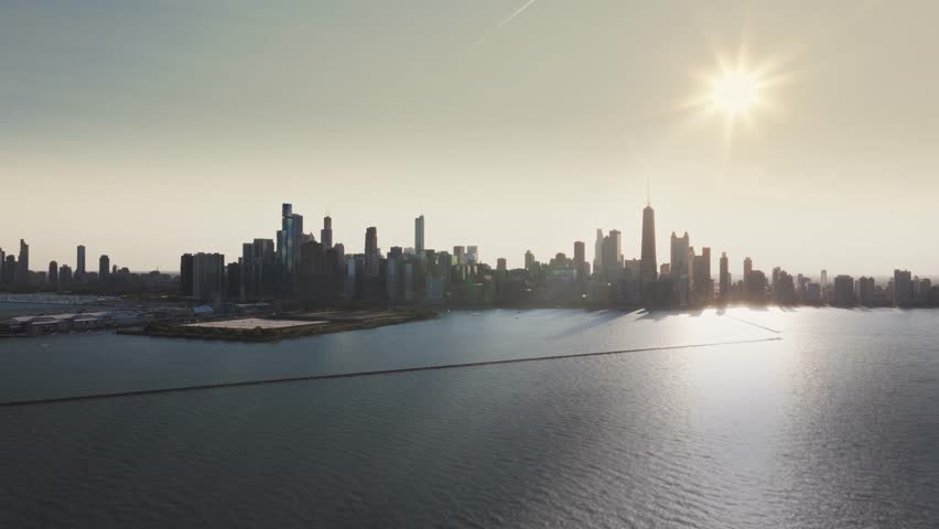 A wide, cinematic drone shot far over Lake Michigan, slowly rotating from left to right, as the setting sun casts a warm glow over the sprawling metropolis and the urban landscape of Chicago, IL.