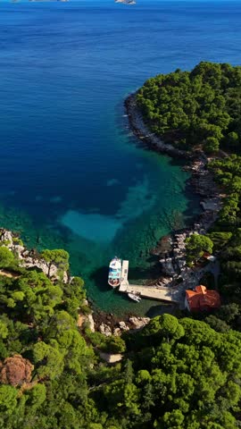 Aerial close-up drone pans right over Lokrum Island’s port, capturing docked boats, surrounding green trees, and the clear blue Adriatic Sea with rocky shores framing the island’s natural harbor.
