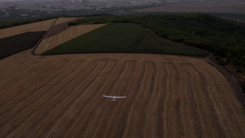 Aerial chasing shot of fixed wing drone flying above agricultural fields towards city of Chisinau at sunset