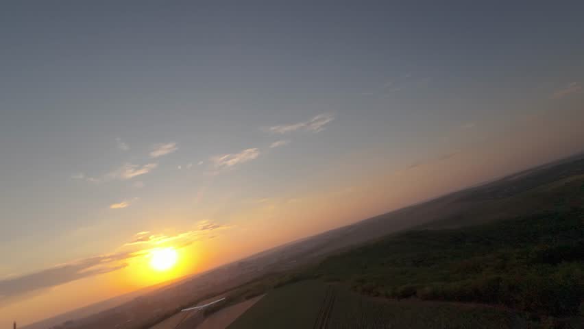 Aerial FPV chasing shot of fixed wing drone flying over fields at sunset with dramatic tilt horizon