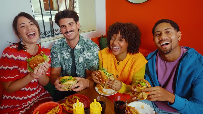 Multiethnic group of four happy friends sitting at a diner table, sharing a meal of burgers and fries while laughing and enjoying each other