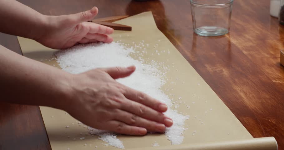 Close up of chef preparing coarse sea salt for curing salmon, spreading and leveling salt on parchment paper to make a bed for fish salting, traditional cooking process, homemade seafood preparation