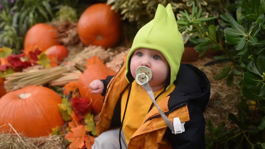 A baby boy in a jacket, hat, and jeans sits among pumpkins outside	