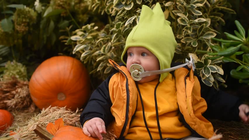 A baby boy in a jacket, hat, and jeans sits among pumpkins outside	
