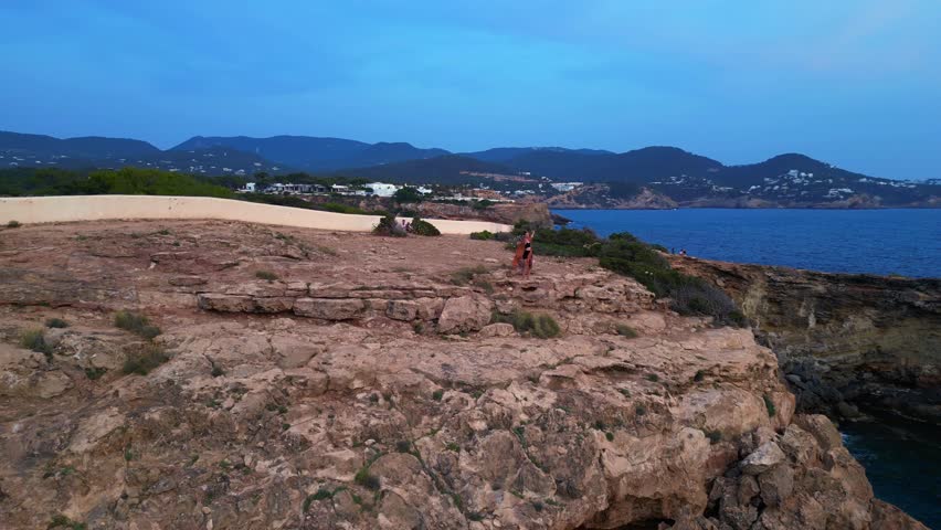 dancing Woman expressing freedom and movement by the sea at a beautiful travel destination Cala Llentia Ibiza. Lovely aerial view flight circle drone footage