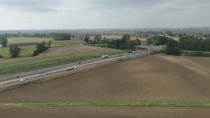 Static drone shot showing cars and trucks traveling along the A21 motorway near Piacenza, Italy, flanked by agricultural fields and agricultural fields, some freshly plowed and others cultivated